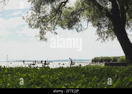 Einfacher Nachmittag am Ufer des Plattensees in Ungarn. Gras am Ufer im Vordergrund mit Angelruten und Wasseraktivitäten im Hintergrund. Stockfoto