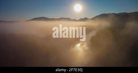Luftdrohnen-Panorama von Fluss und Nebel über nebligen Ebenen mit Hügeln, die während der frühen Sonnenaufgangsstunden aus dem Nebel perren. Am frühen Morgen auf dem Fluss und auf dem Fiel Stockfoto
