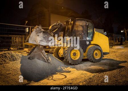 Baustelle an einer Stadtstraße. Ein kleiner, gelber Bagger oder Bobcat, der nachts auf einer Baustelle geparkt hat. Häuser im Hintergrund Stockfoto