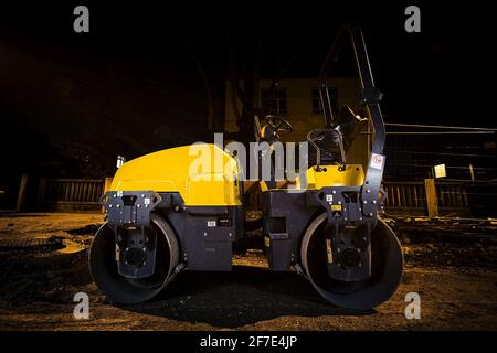 Eine gelbe Asphalt- oder Sandwalze auf einer Vorstadtstraße in der Nacht während der Renovierungsarbeiten. Seitenansicht einer Asphaltwalze. Stockfoto