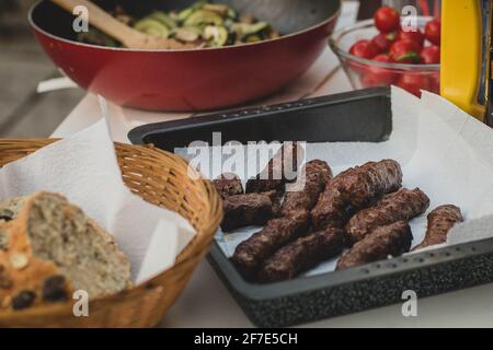 Hausgemachte Cevapcici Mahlzeit auf einem Picknick. Cevapcici in einem Metallbehälter mit Papiertuch, serviert mit Brotkorb und verschiedenen Salaten umgeben. Stockfoto