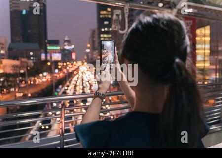 Rückansicht einer jungen, nicht erkennbaren Frau, die auf einer Fußgängerbrücke steht und dabei mitnimmt Bild der Abendansicht der Stadtstraße mit schönem Himmel Anzeigen Stockfoto