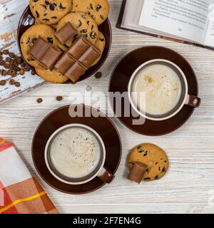 Zwei Tassen Kaffee mit Schokoladenkeksen und Schokolade auf dem Hintergrund eines offenen Buches. Stockfoto
