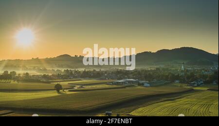 Schönes Morgenpanorama eines Dorfes oder einer Stadt von Sankt Georgen in Attergau, Österreich. Sichtbare Sonne mit Strahlen, die über den Häusern und grünen Wiesen aufgehen. Stockfoto