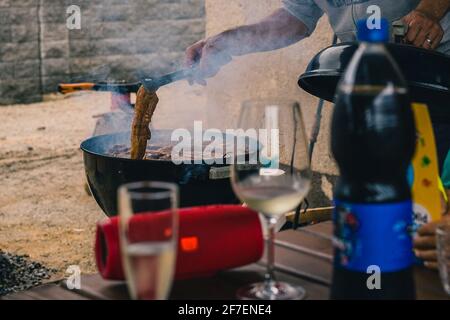Mann, der einen Grill oder einen Holzkohlegrill öffnet, um Kebabs oder Ersatzrippen beim Grillen auf Wärme zu drehen. Rauch vom Grill sichtbar Stockfoto