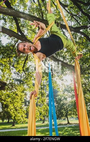 Luftseidenkünstler, der an einem sonnigen Tag im Tivoli Park in Ljubljana Bewegungen an Seilen an Bäumen aufführt. Stockfoto