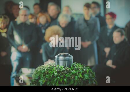 Eine Metallurne mit Asche einer Toten auf einer Beerdigung, mit Menschen, die im Hintergrund bei einer Gedenkfeier trauern. Trauriger trauernder Moment am Ende von Stockfoto