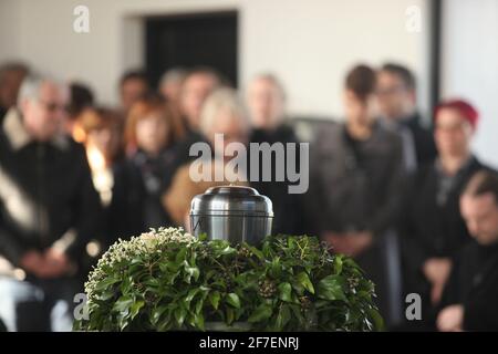 Eine Metallurne mit Asche einer Toten auf einer Beerdigung, mit Menschen, die im Hintergrund bei einer Gedenkfeier trauern. Trauriger trauernder Moment am Ende von Stockfoto