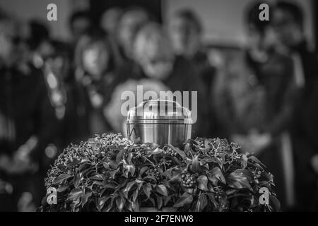 Eine Metallurne mit Asche einer Toten auf einer Beerdigung, mit Menschen, die im Hintergrund bei einer Gedenkfeier trauern. Trauriger trauernder Moment am Ende von Stockfoto