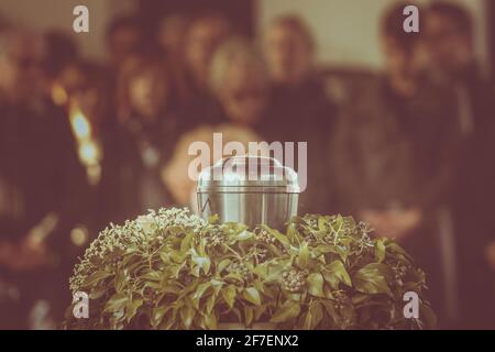 Eine Metallurne mit Asche einer Toten auf einer Beerdigung, mit Menschen, die im Hintergrund bei einer Gedenkfeier trauern. Trauriger trauernder Moment am Ende von Stockfoto