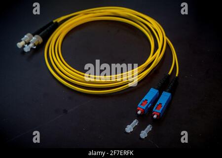 Auf einem grauen Boden liegt eine kurze Rolle aus einem Paar gelber optischer Kabel mit quadratischen Steckverbindern auf einer Seite und rund auf der anderen Seite. Stockfoto