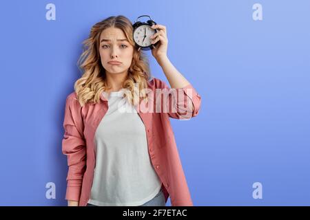 Müde Frau mit Wecker am Kopf befestigt, junge Studentin in Freizeitkleidung mit frustriertem Gesicht Blick auf die Kamera, wollen nicht zur Universität gehen. Isoliert Stockfoto