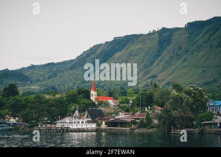 Vulkanische Insel Samosir am Toba-See, auf Nordsumatra, sichtbare typische Häuser und christliche Kirche auf der Insel an einem bewölkten Tag. Stockfoto