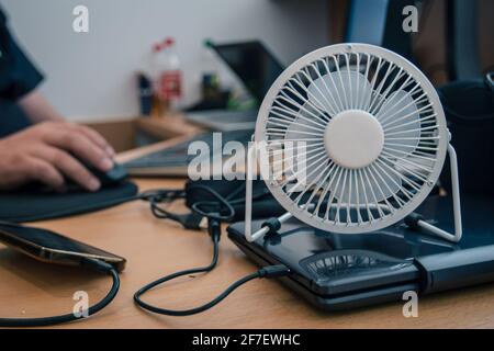 Ein kleiner, USB-betriebener Desktop-Lüfter oder -Ventilator zum Kühlen des Ohrhörers auf einem Schreibtisch mit Desktop-Computer. Hände einer Person, die im Hintergrund arbeitet Stockfoto