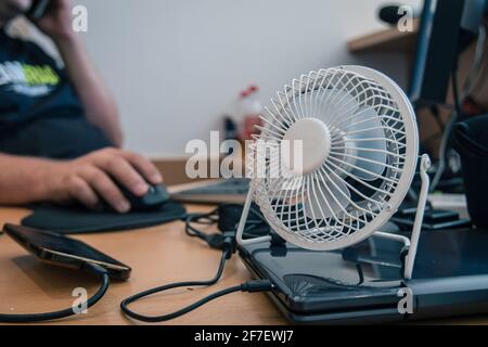 Ein kleiner, USB-betriebener Desktop-Lüfter oder -Ventilator zum Kühlen des Ohrhörers auf einem Schreibtisch mit Desktop-Computer. Hände einer Person, die im Hintergrund arbeitet Stockfoto