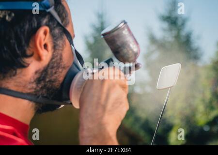 Amateur-Maler in roten Pullover und Gesichtsmaske mit einer Spritzpistole, um ein Kunststoffobjekt oder Teil in einem Hausgarten zu malen. Rückansicht eines Malers mit einem kleinen Stockfoto