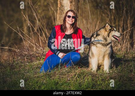 Ein Mädchen mit einem Lächeln kniet sich im Frühjahr an einer Leine auf einem Feld mit Bäumen zu einem sibirischen Husky-Hund nieder. Das Mädchen trägt ein rotes und blaues Leder Stockfoto