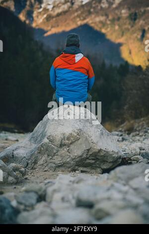 Blick auf einen modernen Hipster im Freien, der auf einem Felsen neben einem Fluss sitzt und nachdenkt. Ein denkender Mann in der freien Natur. Auf der Suche nach Einsamkeit Stockfoto