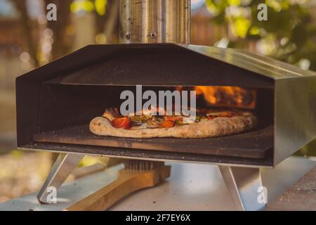 Hausgemachte Pizza wird in einem tragbaren Aluminium-Backofen für Pizza eingesetzt. Köstliche Pizza wird in einem Ofen gebacken, sichtbares Feuer auf der Rückseite des Furnás Stockfoto