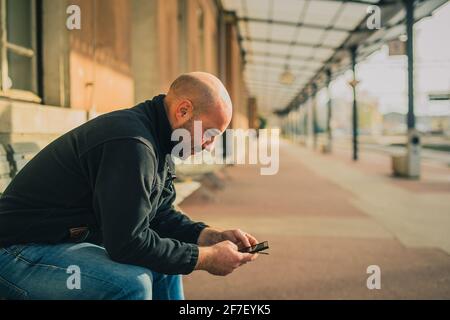 Seitenansicht eines jungen kahlen Mannes, der auf einer Bank sitzt und ein Telefon benutzt und raucht, während er auf einen Zug auf einem älteren Vintage-Bahnhof wartet. Stockfoto