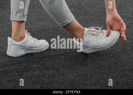 Die junge Frau beugte sich und berührte ihre Füße mit weißen Sneakern mit einer Hand als Teil einer Aufwärm-Routine in Innenräumen. Detail auf den Schuhen, unbekannt woma Stockfoto