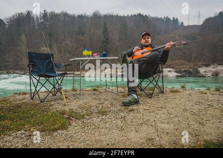 Mann in der freien Natur, der auf einem Stuhl sitzt und Gitarre spielt. Kalter Camping neben einem Fluss und einem Wald. Genießen Sie ein wildes Zelten in Stockfoto