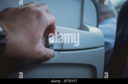 Man öffnet den Knopf zum Lösen der Tischschale an der Rückenlehne eines Flugzeugsitzes. Sichtbares Tablett in verstauter Position mit der Hand einer Person, die den Knopf dreht. Stockfoto