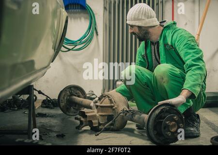 Ein junger Mann entfernt eine große Hinterachse aus einem alten Muskelwagen von der Unterseite des Autos. Auto Reparatur oder Restaurierung zu Hause. Stockfoto