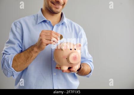 Unbekannter Mann, der auf grauem Hintergrund steht, legt eine Münze in Form eines Schweins in ein Sparschwein. Stockfoto