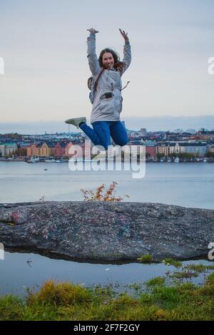 Die junge Frau springt auf den Aussichtspunkt Skinnarviksberget, weit über dem Stockholmer Schloss. Das Stadtbild von stockholm ist im Hintergrund zu sehen. Stockfoto