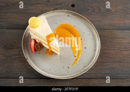 Blick von oben auf leckeren orangen und weißen Kuchen auf dem Teller, isoliert auf einem Holztisch. Köstliche hausgemachte Dessertscheibe mit Beeren, Makronen-Plätzchen und mit Aprikosenpüree dekoriert. Stockfoto