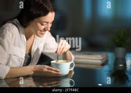 Glückliche Frau, die Kaffee rührt, sitzt im Wohnzimmer Die Nacht zu Hause Stockfoto