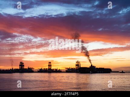 Las Palmas, Gran Canaria, Kanarische Inseln, Spanien. April 2021. Ein Containerschiff verdunkelt den schwarzen Rauch, während der Himmel mit Farbe explodiert, während die Sonne hinter Bohrschiffen im Hafen von Las Palmas auf Gran Canaria aufgeht. Im Hafen befinden sich rund zehn Bohrschiffe und Bohrinseln, die seit mehr als einem Jahr stillstehen. Quelle: Alan Dawson/Alamy Live News. Stockfoto