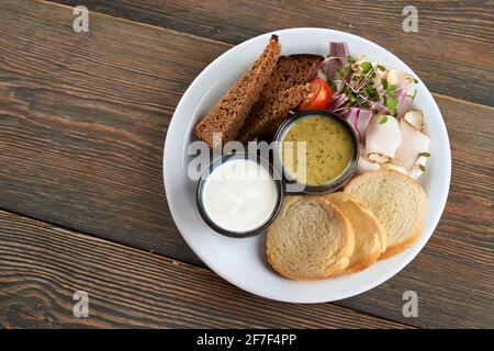 Die weißen und dunklen Brotscheiben werden von oben mit Sprossen, Gemüse und Schweinefett auf dem Teller serviert. Leckerer Snack mit saurer Rahm und grüner Sauce im Restaurant. Konzept der traditionellen ukrainischen Küche. Stockfoto