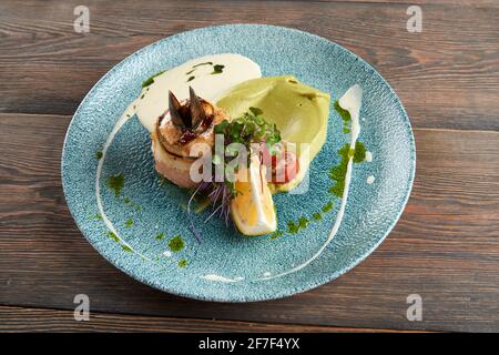 Draufsicht auf große gebackene Brötchen mit Fisch, Käse und Muscheln in schwarzer Schale, serviert mit Avocado-Purre, Zitrone, frischem Sprossen, Kirschtomaten und saurer Creme auf einer schönen blauen Keramikplatte. Stockfoto