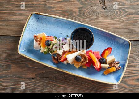 Von oben Blick auf lange blaue Platte mit Früchten, Beeren, Nüssen und Sprossen auf verschiedenen Käsescheiben, Glas dunkle Sauce in der Nähe. Leckere Vorspeise, zubereitet im Luxusrestaurant. Konzept der Küche. Stockfoto