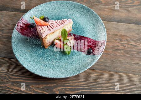 Von oben Blick auf den traditionellen Käsekuchen mit Heidelbeeren und Preiselbeeren auf dem Teller mit violettem Pinselstrich Dekoration serviert. Leckeres Essen auf einem Holztisch im Restaurant. Konzept von Dessert, Servieren. Stockfoto