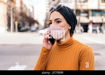 Ausdrucksstarke junge muslimische Frau, die mit ihrem Mobiltelefon auf der Straße eine schützende Gesichtsmaske trägt. Stockfoto