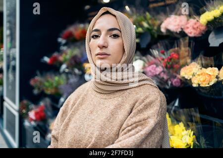 Schöne junge muslimische Frau, die gegen den Blumenladen steht und die Kamera anschaut. Stockfoto