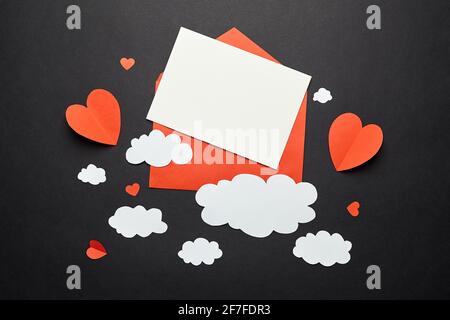 Grußkarten-Mockup mit Papierdekorationen auf schwarzem Hintergrund. Leere Grußkarte oder Einladung und weiße Papierwolken, roter Umschlag und Herzen Stockfoto