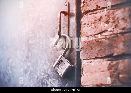 Am Griff einer eisernen Tür hängt ein altes, offenes Metallschloss, das in einem Backsteingebäude steht und von Sonnenlicht beleuchtet wird. Ein offenes Geheimnis. Stockfoto
