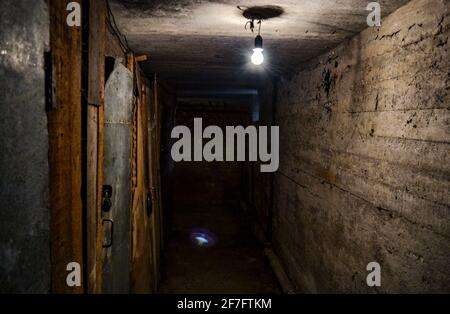 Ein schrecklicher dunkler Betonkorridor im Keller mit verschlossenen Türen an den Seiten, beleuchtet von einer einzigen Glühbirne, die von der niedrigen Decke hängt. Stockfoto