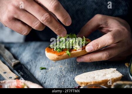 Nahaufnahme eines jungen kaukasischen Mannes, der mit gehacktem Salat eine vegane Vorspeise mit einem halbierten gerösteten Brotbrötchen, das mit einer Tomate und einer Mandel bedeckt ist, zubereitet Stockfoto