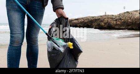 Ein Mann sammelt einige Abfälle, wie Dosen, Flaschen oder Taschen, aus dem Sand eines einsamen Strandes, um die natürliche Umgebung zu reinigen Stockfoto