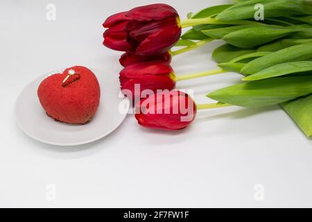 Ein Bouquet aus roten Tulpen neben einem Teller mit einem herzförmigen Kuchen, in dem ein Goldring mit einem Diamanten. Stockfoto