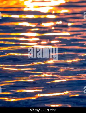 Meereswellen-Textur mit Sonne- oder Sonnenaufgangslicht, das auf dem Wasser scheint. Sonnenstrahlen beleuchten und reflektieren das blaue Meerwasser mit warmem orangefarbenem Licht Stockfoto