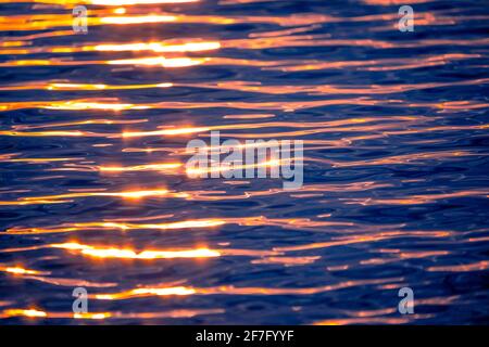 Meereswellen-Textur mit Sonne- oder Sonnenaufgangslicht, das auf dem Wasser scheint. Sonnenstrahlen beleuchten und reflektieren das blaue Meerwasser mit warmem orangefarbenem Licht Stockfoto