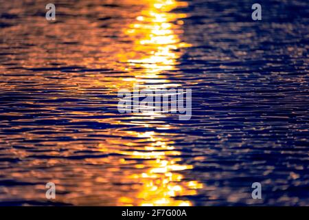 Meereswellen-Textur mit Sonne- oder Sonnenaufgangslicht, das auf dem Wasser reflektiert wird. Sonnenstrahlen beleuchten das blaue Meerwasser mit warmem orangefarbenem Licht. Nahaufnahme. Stockfoto