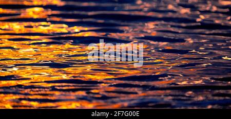 Meereswellen-Textur mit Sonne- oder Sonnenaufgangslicht, das auf dem Wasser reflektiert wird. Sonnenstrahlen leuchten auf dem blauen Meerwasser mit warmem orangefarbenem Sonnenlicht. Artisti Stockfoto