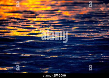 Meereswellen-Textur mit Sonne- oder Sonnenaufgangslicht, das auf dem Wasser reflektiert wird. Sonnenstrahlen leuchten auf dem blauen Meerwasser mit warmem orangefarbenem Sonnenlicht. Artisti Stockfoto